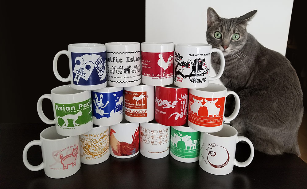 A gray cat sits next to 15 mugs from previous banquets stacked on top of one another.